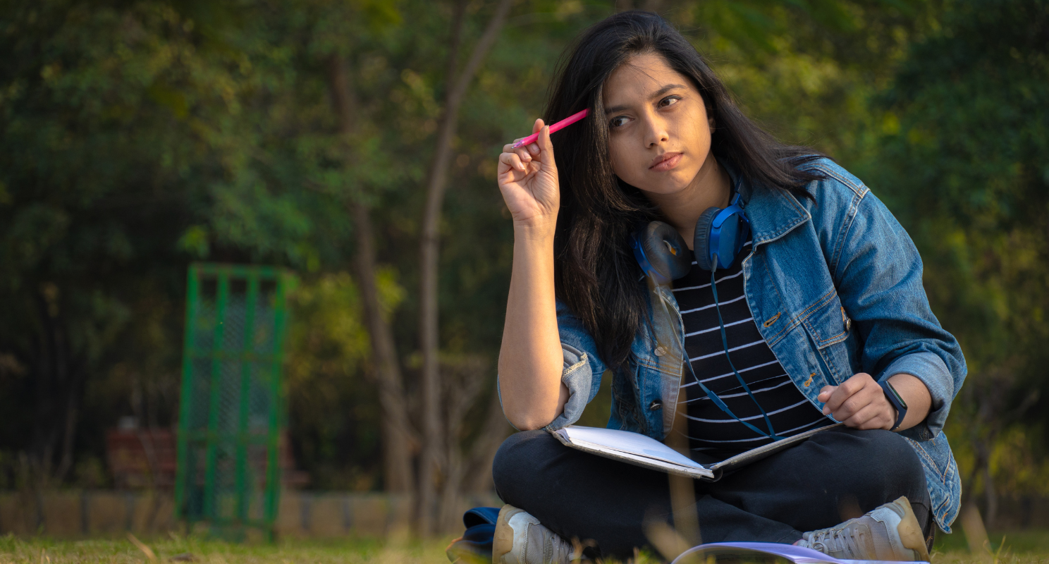 Girl student sat cross-legged on the grass with notebook and pen thinking