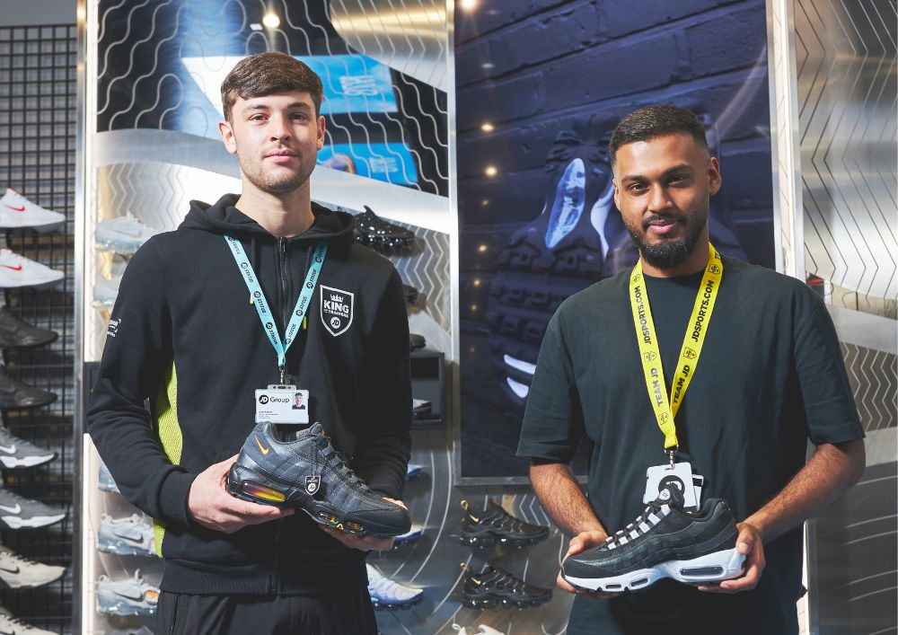 Two people stand in a shoe store, each holding a sneaker. Display shelves with various footwear are visible behind them.