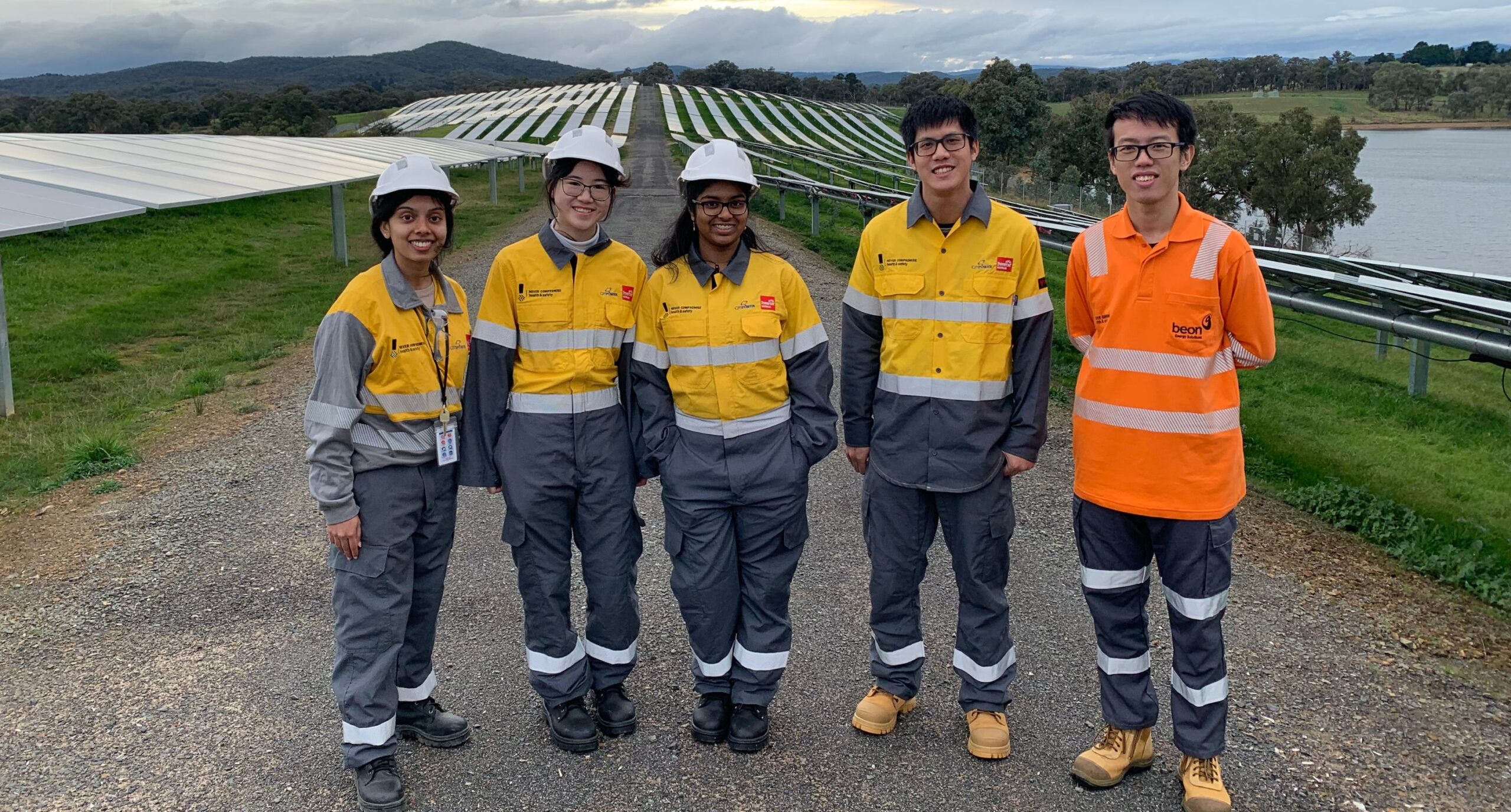 Four individuals in bright orange and yellow work attire stand proudly beside a solar farm, showcasing renewable energy efforts.