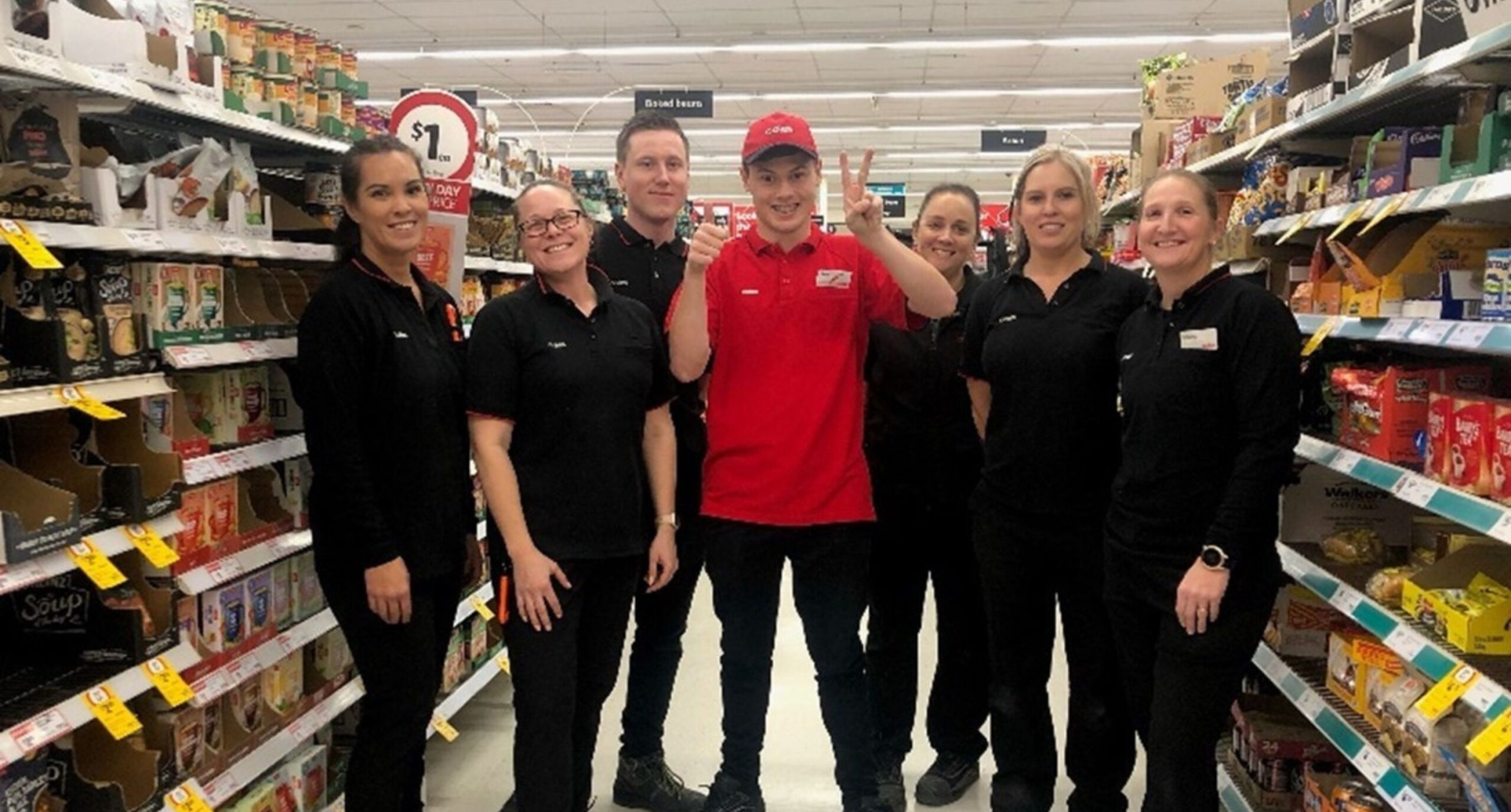 A diverse group of Coles employees chatting and shopping in a brightly lit supermarket aisle filled with various products.