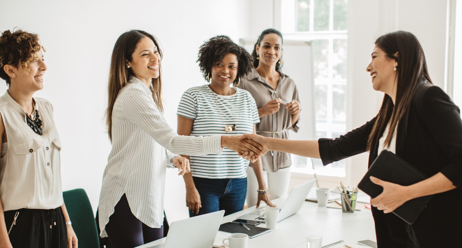 four women stand across the table from their boss and one shakes the bosses hand