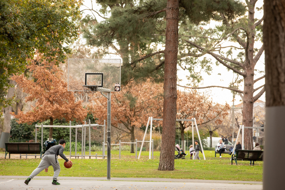 Young boy playing basket ball in a public park with grass and swings in background