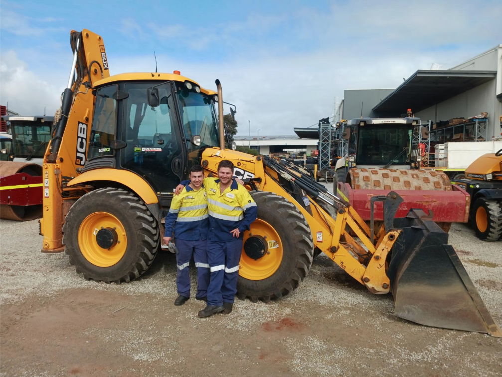Brothers, Mathew and Kristian smiling with arms over each other's shoulders, with a large JCB digger in the background