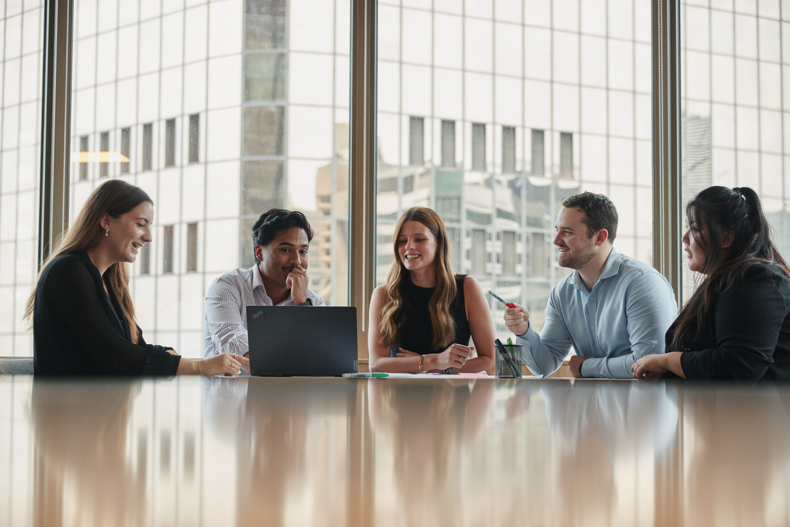 Colleagues sat across a long table, with tall city buildings in view out of the window